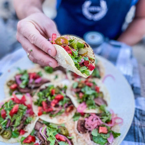 A person holds up a taco loaded with fresh toppings and a sprinkle of Gustus Vitae Ultimate Gourmet BBQ Collection - 6 Tins seasoning, above a plate of similar vibrant tacos. The background is blurred.