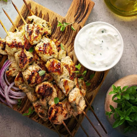 Grilled chicken skewers on a wooden board, seasoned with Gustus Vitaes Ultimate Gourmet BBQ Collection - 6 Tins, served with sliced red onions, fresh herbs, creamy dipping sauce, olive oil, and a small herb plant nearby.