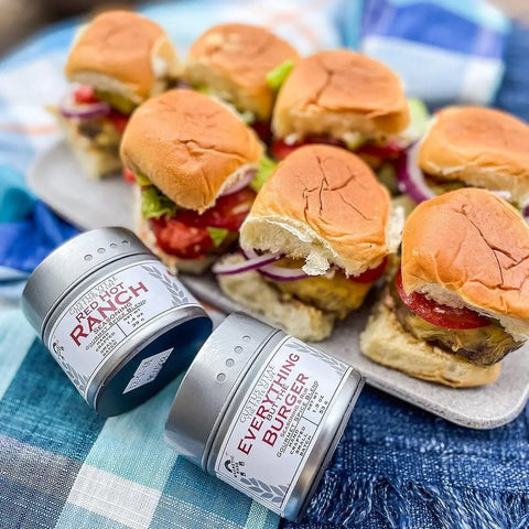 A tray of mini burgers sits on a checkered cloth—ideal for a BBQ. In front, two tins from Gustus Vitaes Gourmet Backyard BBQ Burgers & Fries Seasoning Set - Six Pack are displayed.