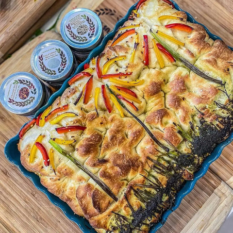 The Luxury Beginner Cook - 4 Tins by Gustus Vitae features premium seasonings in magnetic tins, displayed beside a focaccia topped with vibrant peppers and herbs arranged like flowers on a wooden surface.