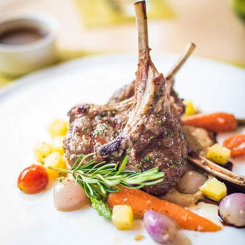 A plate of grilled lamb chops seasoned with Gustus Vitaes Luxury Beginner Cook - 4 Tins, garnished with rosemary and served alongside baby carrots, cherry tomatoes, pearl onions, diced yellow veggies, and a savory sauce.