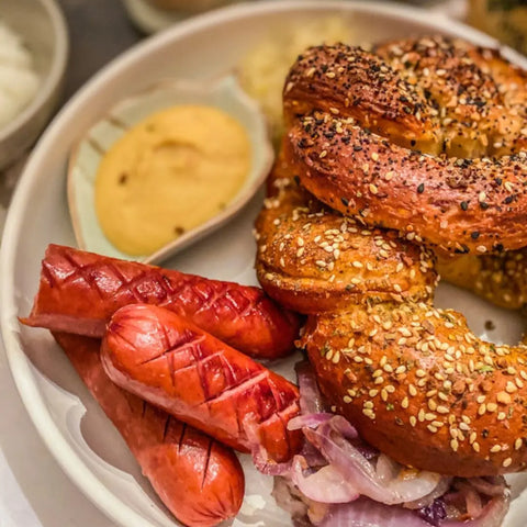 Served on the side: two grilled sausages with Gustus Vitae’s premium seasonings from the Luxury Beginner Cook - 4 Tins, sliced onions, two seeded twisted pretzels, and creamy yellow mustard.