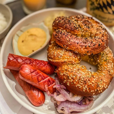 A white dish holds a sesame seed pretzel, two grilled sausages, sautéed onions seasoned with Gustus Vitae’s Breakfast & Brunch Seasoning Set, gourmet spices, sauerkraut, and mustard dip—all neatly arranged.