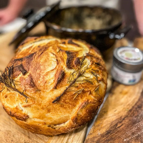 A golden-brown artisan loaf topped with rosemary rests on a wooden board, while Gustus Vitae’s Breakfast & Brunch Seasoning Set and a black cast iron pot add gourmet flair to the scene.