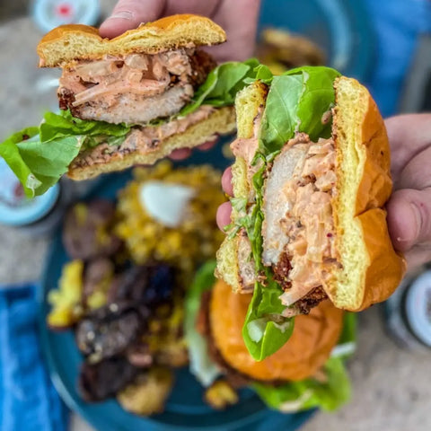 A close-up of hands holding a fried chicken sandwich with lettuce and creamy sauce, cut in half to reveal fillings enhanced by Gustus Vitae’s Deluxe Chicken BBQ Seasonings Collection – 3 Pack. Grilled meats, corn, and another sandwich appear in the background.