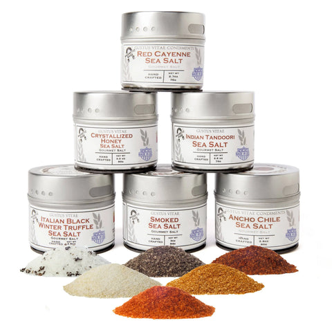 The Gustus Vitae Finishing Sea Salts Collection features 6 tins, including smoked and truffle sea salts, stacked pyramid-style with small piles of each colorful blend shown in front on a white background.