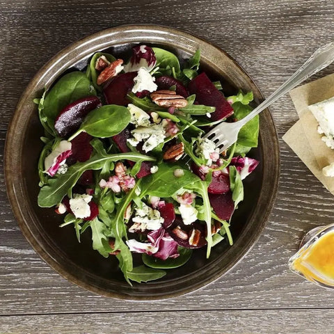 A bowl of fresh salad with arugula, spinach, sliced beets, blue cheese, and pecans on a wooden table is finished with Gustus Vitae’s Finishing Sea Salts Collection for extra flavor. Cheese and dressing are served on the side.