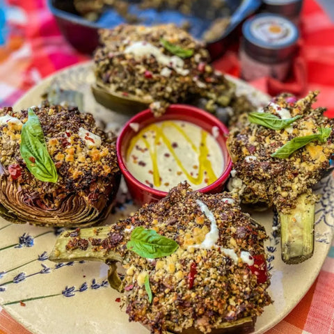 A plate with four stuffed artichokes, garnished with Gustus Vitae’s Gourmet French Fry Seasoning Set - Six Pack, surrounds a bowl of creamy dipping sauce. Colorful tablecloth and spice jars appear in the background.