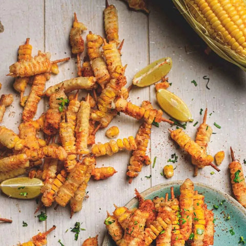 Spiral-cut grilled corn skewers, seasoned with Gustus Vitaes Gourmet French Fry Seasoning Set - Six Pack, are arranged on a light wooden surface with lime wedges, herbs, and a nearby basket of corn on the cob.