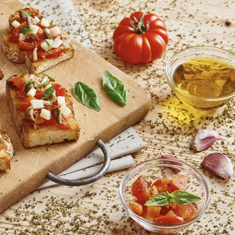Slices of bruschetta topped with diced tomatoes, cheese, basil, and Gustus Vitae’s Garlic Lovers Collection sea salt sit on a wooden board next to a whole tomato, bowls of olive oil and tomatoes, garlic cloves, and scattered herbs.