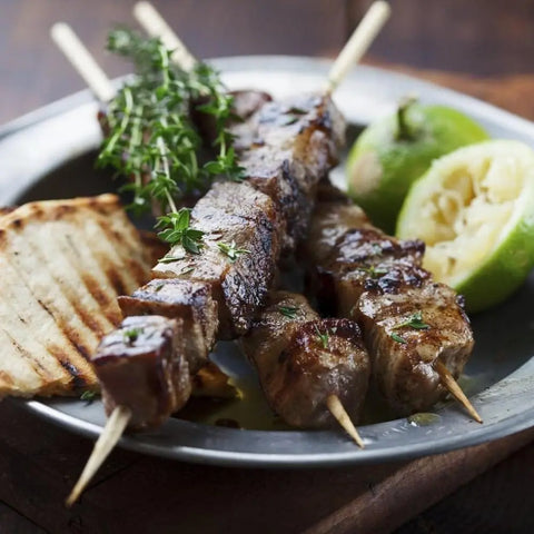 Experience the Taste of Greece with Gustus Vitaes Garlic Lovers Collection - 3 Tins, featuring grilled meat skewers seasoned with garlic pepper and herbs, served alongside grilled bread and lime for a flavorful feast.