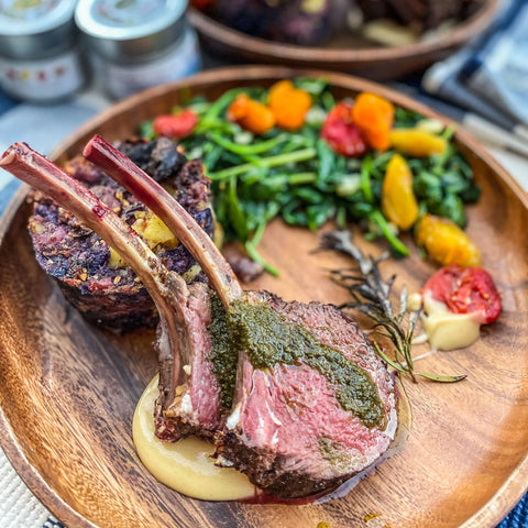 A wooden plate with two lamb chops seasoned with Gustus Vitae Pantry Essentials Set - 4 Tins, topped with green herb sauce on creamy puree, roasted vegetables, leafy greens, and a side of baked potato or stuffing.