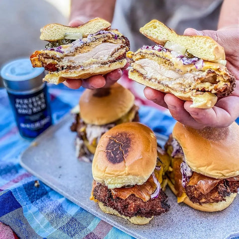 A person holds a halved fried chicken sandwich with pickles, onions, and sauce above a platter of similar sandwiches on a checkered cloth. Nearby is Gustus Vitaes Perfect for Poultry Complete 6 Pack Collection of gourmet seasonings and rubs.