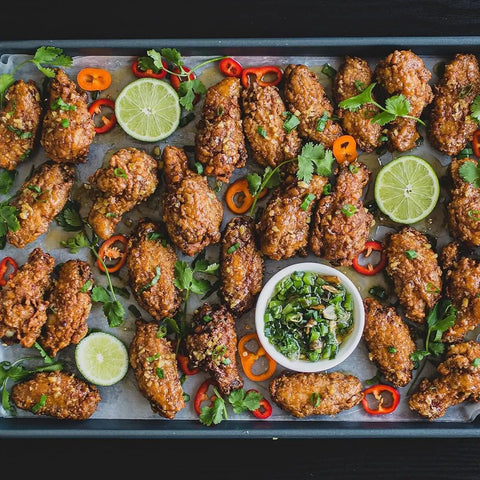 A baking tray of glazed chicken wings seasoned with Gustus Vitae’s Perfect for Poultry 6 Pack Collection, garnished with cilantro, sliced red and orange chilis, and lime slices, served with a dipping sauce in the center.