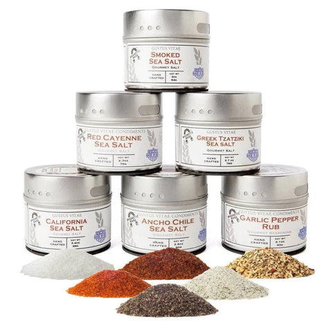 Six tins from the Gustus Vitae Gourmet Finishing Sea Salts and Rubs Collection are stacked in a pyramid, each showcasing a unique flavor, with small piles of these vibrant seasonings displayed in front.
