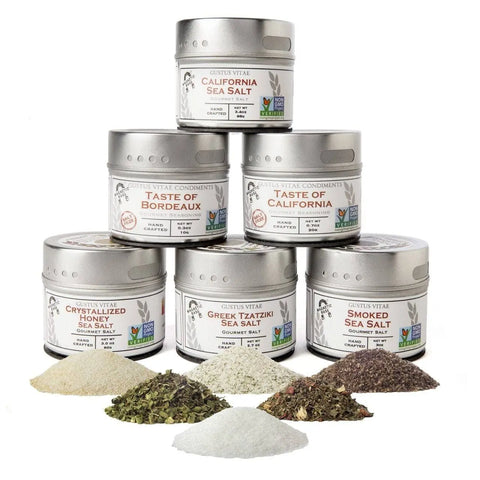 Six tins from the Gustus Vitae Picnic Basket Collection are stacked in a pyramid, each with a vibrant pile of artisan seasoning in front, highlighting the collection’s colorful and textured gourmet salts.