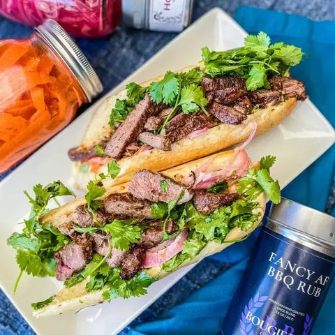 A plate holds two baguette sandwiches with grilled beef, cilantro, carrots, and pickled onions. Nearby are jars of pickled veggies, a Gustus Vitae Fancy AF BBQ Rub tin, and the Picnic Basket Collection - 6 Tins from Gustus Vitae.