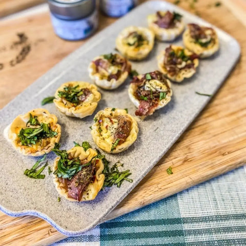 The Gustus Vitae Picnic Basket Collection - 6 Tins features savory baked phyllo cups with artisan seasonings and herbs, artfully arranged on a stone platter atop a wooden board accented by a blue plaid cloth.