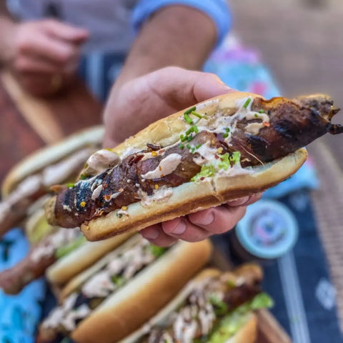 A hand holds a grilled sausage in a bun, topped with creamy sauce, chives, and Gustus Vitae artisan seasonings from the Picnic Basket Collection - 6 Tins. More similarly topped hot dogs appear in the blurred background.