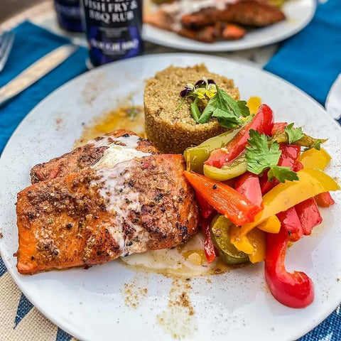 A white dish featuring grilled salmon seasoned with Gustus Vitaes Superb for Seafood 6 Pack Collection, topped with creamy sauce, quinoa with herbs, and sautéed red, yellow, and green bell peppers.