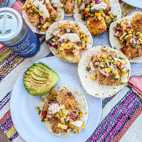 A plate of fish tacos with corn salsa and creamy sauce, seasoned with Gustus Vitaes Superb for Seafood 6 Pack gourmet rubs, sits by sliced avocado. More tacos, Pacifico Claro beer, and a vibrant woven tablecloth appear in the background.