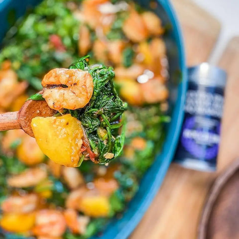 A wooden spoon lifts shrimp, leafy greens, and yellow squash over a vibrant blue dish of saucy vegetables; in the background, Gustus Vitaes Superb for Seafood: Complete 6 Pack gourmet seasonings and rubs add a touch of culinary flair.