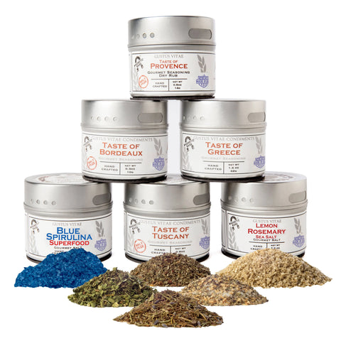 Six tins of Gustus Vitaes Luxury Gourmet Seasoning and Sea Salt Collection are stacked in a pyramid, each featuring a unique flavor. Small piles of assorted herbs, spices, and blue sea salt are arranged in front.