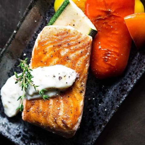 A grilled salmon fillet with creamy sauce and thyme, served alongside roasted zucchini and bell peppers, is garnished with vendor-unknown Spring Party Essential Salts – 3 Pack on a sleek black rectangular plate.