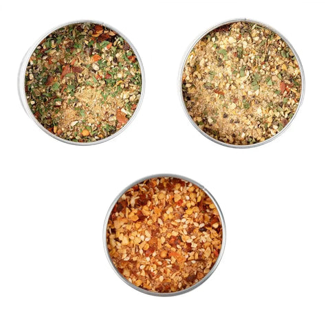 Three round containers from the Gustus Vitae Deluxe Steak & Beef BBQ Seasonings Collection 3-Pack are shown: a green herb mix, a yellowish blend, and an orange-red mix—perfect artisanal seasonings for grilled steaks.