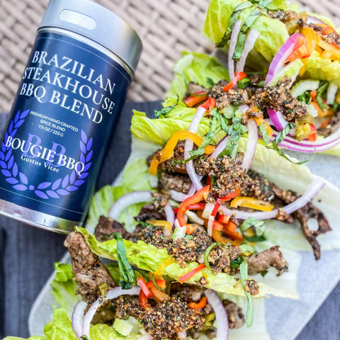 A tin from the Gustus Vitae Deluxe Steak & Beef BBQ Seasonings Collection - 3 Pack sits beside lettuce wraps filled with sliced beef, bell peppers, red onion, and herbs, all arranged on a woven surface.