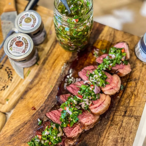 Sliced medium-rare grilled steak with chimichurri sauce on a wooden board, served alongside extra chimichurri and the Gustus Vitae Deluxe Steak & Beef BBQ Seasonings Collection - 3 Pack of gourmet spice blends.