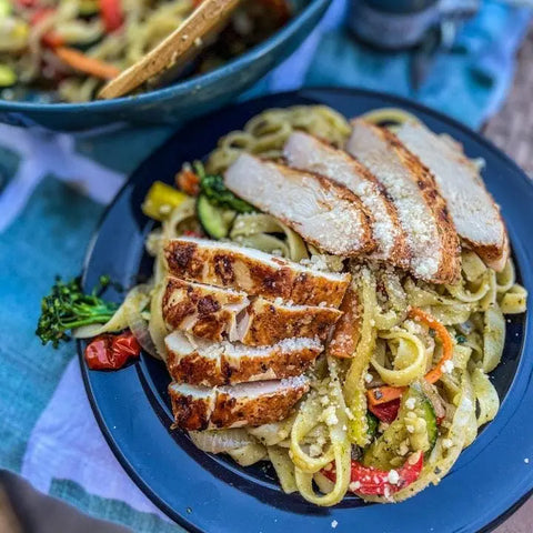 A plate of fettuccine with vegetables and grilled chicken, topped with cheese and Gustus Vitaes Essential Summer Spices & Sea Salts Pantry Upgrade, sits on a blue dish with a fork. Another pasta bowl is in the background.