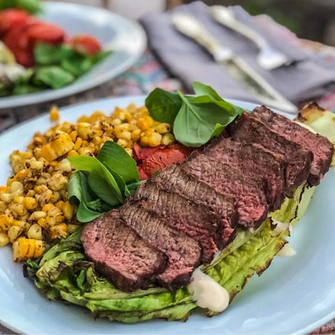 A plate of grilled steak on romaine with roasted corn, spinach, and grilled tomato is enhanced by Gustus Vitae’s Essential Summer Spices & Sea Salts Pantry Upgrade | Set of 4. Silverware and another plate are seen in the background.