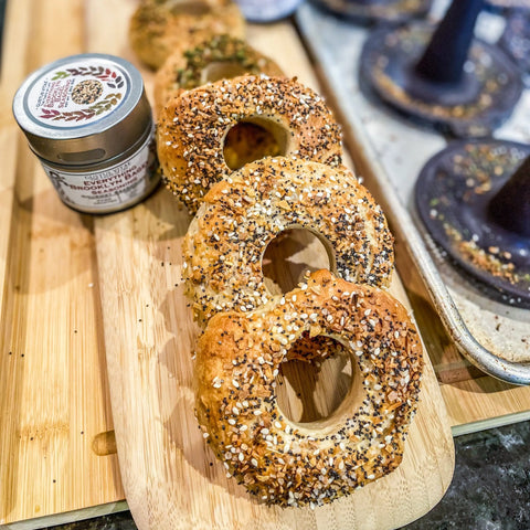 Four everything bagels are arranged on a wooden board beside a tin from the Gustus Vitae Essential Summer Spices & Sea Salts Pantry Upgrade set of 4, with more bagels and baking domes visible in the background.