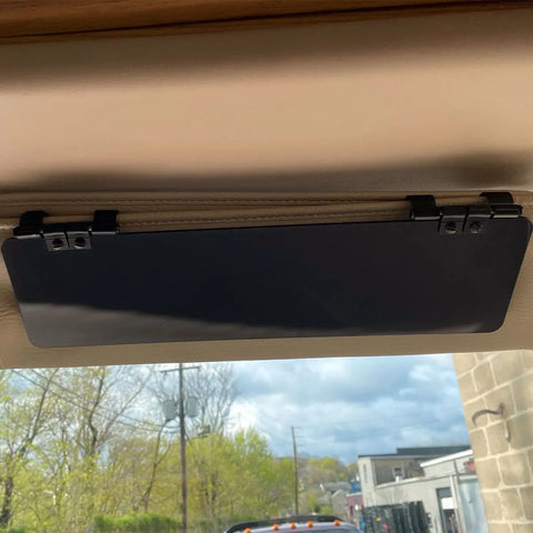 A Glare Guard Gray Polarized Truck Sun Visor Extender is mounted to the car ceiling above the windshield, with trees, utility poles, and buildings visible outside under a cloudy sky.