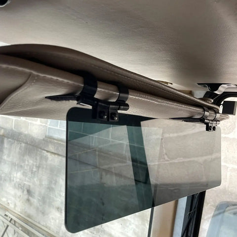 A Glare Guard Gray Polarized Truck Sun Visor Extender is clipped onto a beige car sun visor, viewed from below inside the vehicle, with an exterior stone wall visible through the window.