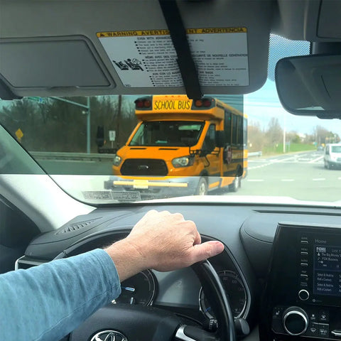 From the driver’s view using the Glare Guard Gray XL MaxView Polarized Car Visor Extender, a car approaches a stopped yellow school bus at an intersection, with the drivers hand on the steering wheel and road signs in sight.