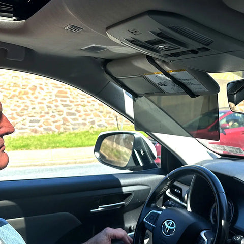 A person drives a Toyota on a sunny day, using the Glare Guard Gray XL MaxView Polarized Car Visor Extender. The car’s interior, steering wheel, part of the driver’s face and hand, plus a stone wall and red car outside are visible.