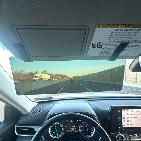 View from inside a car driving at 75 mph, dashboard and speedometer visible. Several cars ahead, trees and a concrete wall on the right. The Glare Guard Gray XL MaxView Polarized Car Visor Extender helps reduce glare for safer driving.