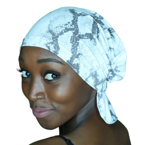 A woman with smooth brown skin wears a white and gray patterned Scarves - Tammy Chemo Beanie® by Chemo Beanies®, looking over her shoulder and smiling softly at the camera against a black background.