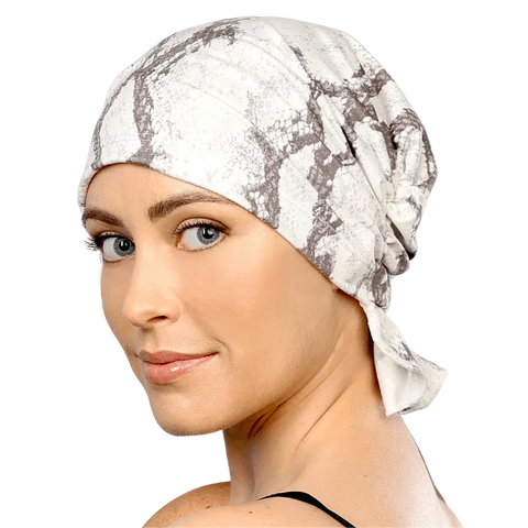 A woman with light skin and blue eyes models a white and gray patterned Scarves - Tammy Chemo Beanies® by Chemo Beanies®, smiling slightly over her shoulder against a black background.