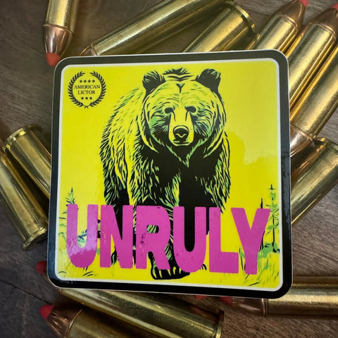 The Sticker: Grizzly - Unruly by American Lictor features a yellow design with a black bear and bold pink UNRULY text, surrounded by .44 magnum bullets with copper tips on a wooden surface.