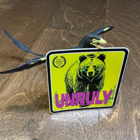 A toy pterosaur stands behind an American Lictor Sticker: Grizzly - Unruly, featuring a bear and laurel, as if guarding an animal deterrent on a wooden floor.