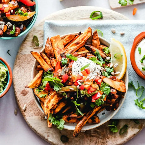 A plate of sweet potato fries with black beans, salsa, sour cream, herbs, seeds, and lemon is served on a wooden board, surrounded by bowls of dips and garnishes featuring Gustus Vitaes Ground Single Ingredient Pepper Collection.