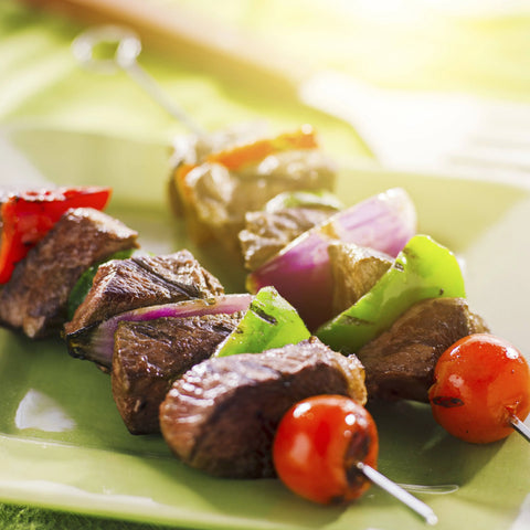Two grilled beef kebabs, seasoned with Gustus Vitaes Ultimate BBQ & Grilling Seasonings 12 Pack Collection, are served with red onion, green bell pepper, and cherry tomatoes on a green plate in warm sunlight.