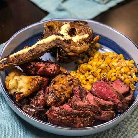 A bowl of sliced steak seasoned with Gustus Vitaes Ultimate BBQ & Grilling Seasonings, roasted potatoes, charred corn, and a grilled artichoke with melted butter sits on a blue cloth napkin.