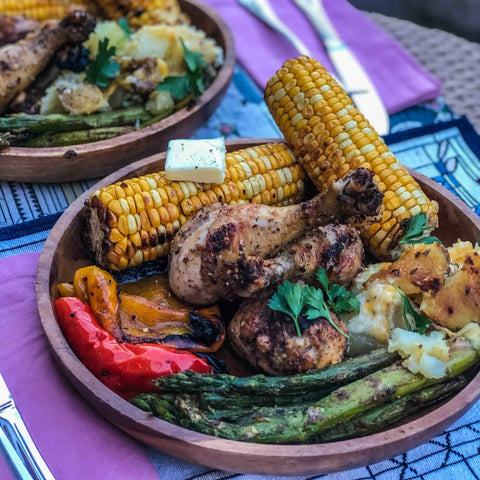 A wooden bowl of grilled chicken drumsticks seasoned with Gustus Vitaes Ultimate BBQ & Grilling Seasonings, corn on the cob, peppers, asparagus, parsley, and mashed potatoes sits on a colorful placemat with cutlery and a purple napkin.