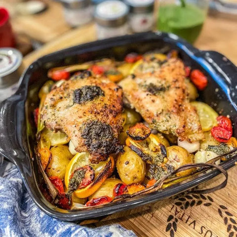A savory baked dish with two chicken thighs seasoned with Gustus Vitaes Slow Cooker Seasonings - 6 Spice Blends Gift Set, atop roasted potatoes, sliced lemons, cherry tomatoes, and herbs in a black square baking dish on a wooden table.