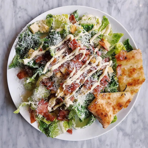 Enjoy a Caesar salad topped with cheese, tomatoes, bacon bits, croutons, and creamy ranch dressing from Gustus Vitae’s All Ranch Everything Collection | Set of 4, served with toasted flatbread on a marble surface.