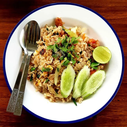 A plate of fried rice made with Gustus Vitae’s Fried Rice Seasoning Collection is garnished with fresh herbs, cucumber slices, and lime, served on a blue-rimmed white plate with fork and spoon atop a wooden table.